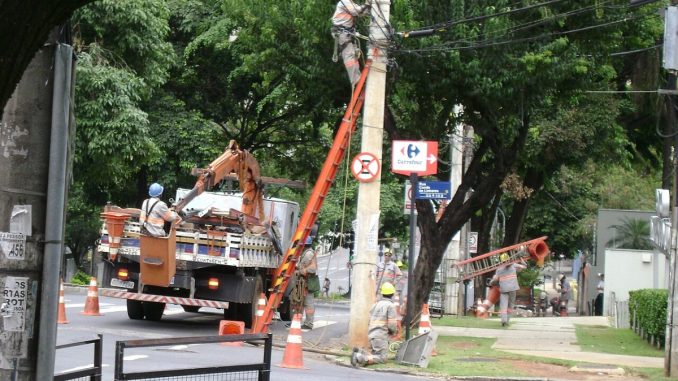 Eletricista no alto de um poste e caminhões com braço mecânico em rua arborizada, durante serviço na rede elétrica
